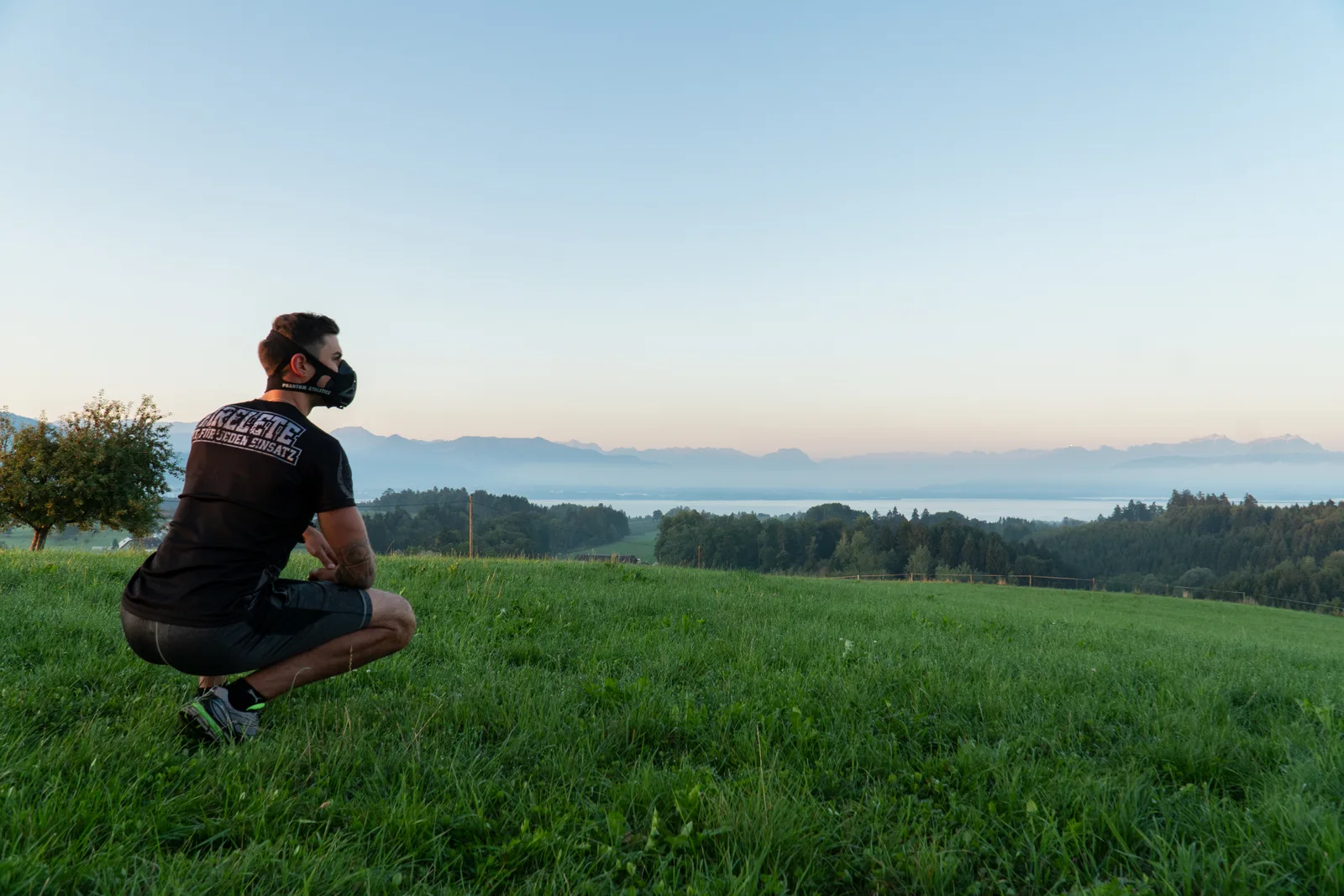 Athlet in Atemschutzmaske beim Fitnesstraining auf Bergwiese mit Seeblick