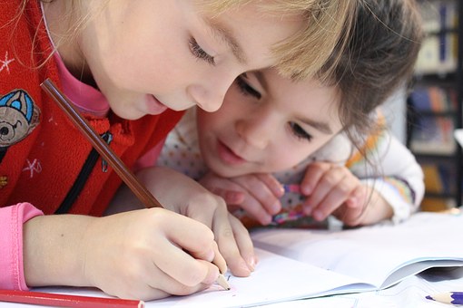 Zwei Kinder schreiben gemeinsam an einem Tisch, eines in rotem Oberteil konzentriert auf Papier blickend