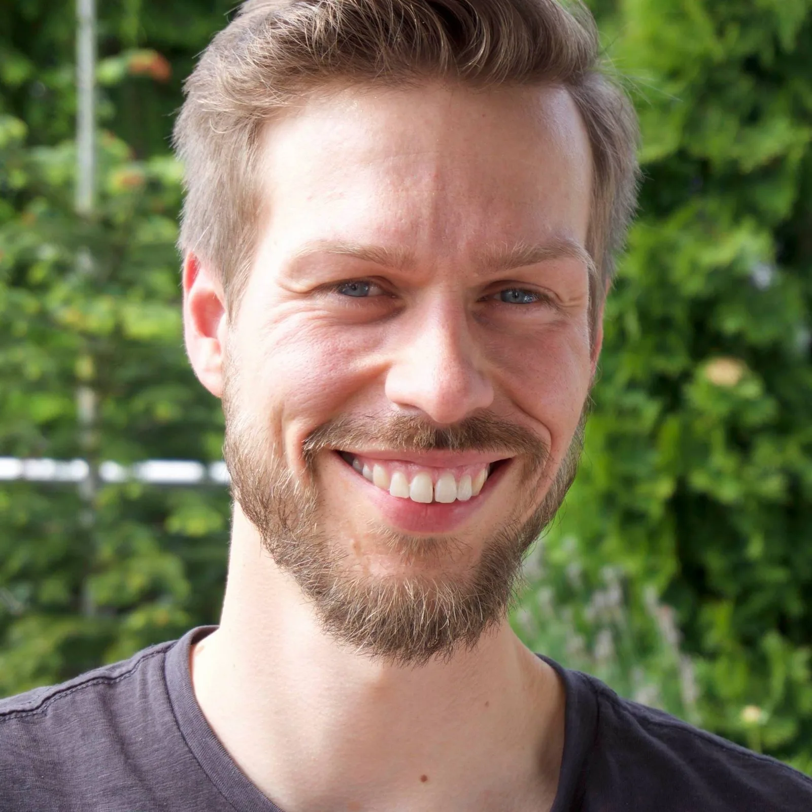 Porträt eines Mannes mit beige-braunem Haar, Vollbart, schwarzem T-Shirt vor grünem Garten-Hintergrund lächelnd