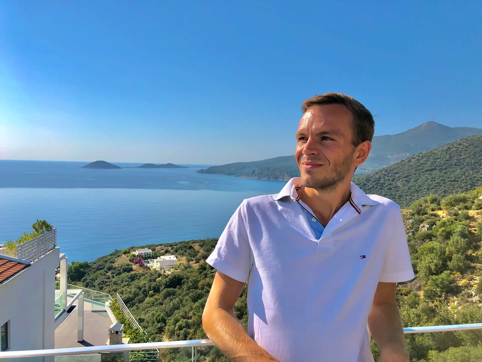 Mann in weißem Poloshirt an Balkon mit Meerblick und Insellandschaft