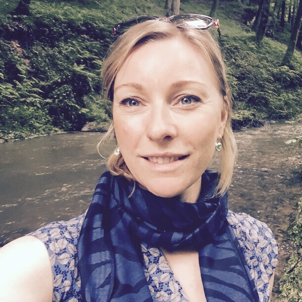 Frau mit blondem Haar und Brille lächelt im Wald, trägt Schal um den Hals