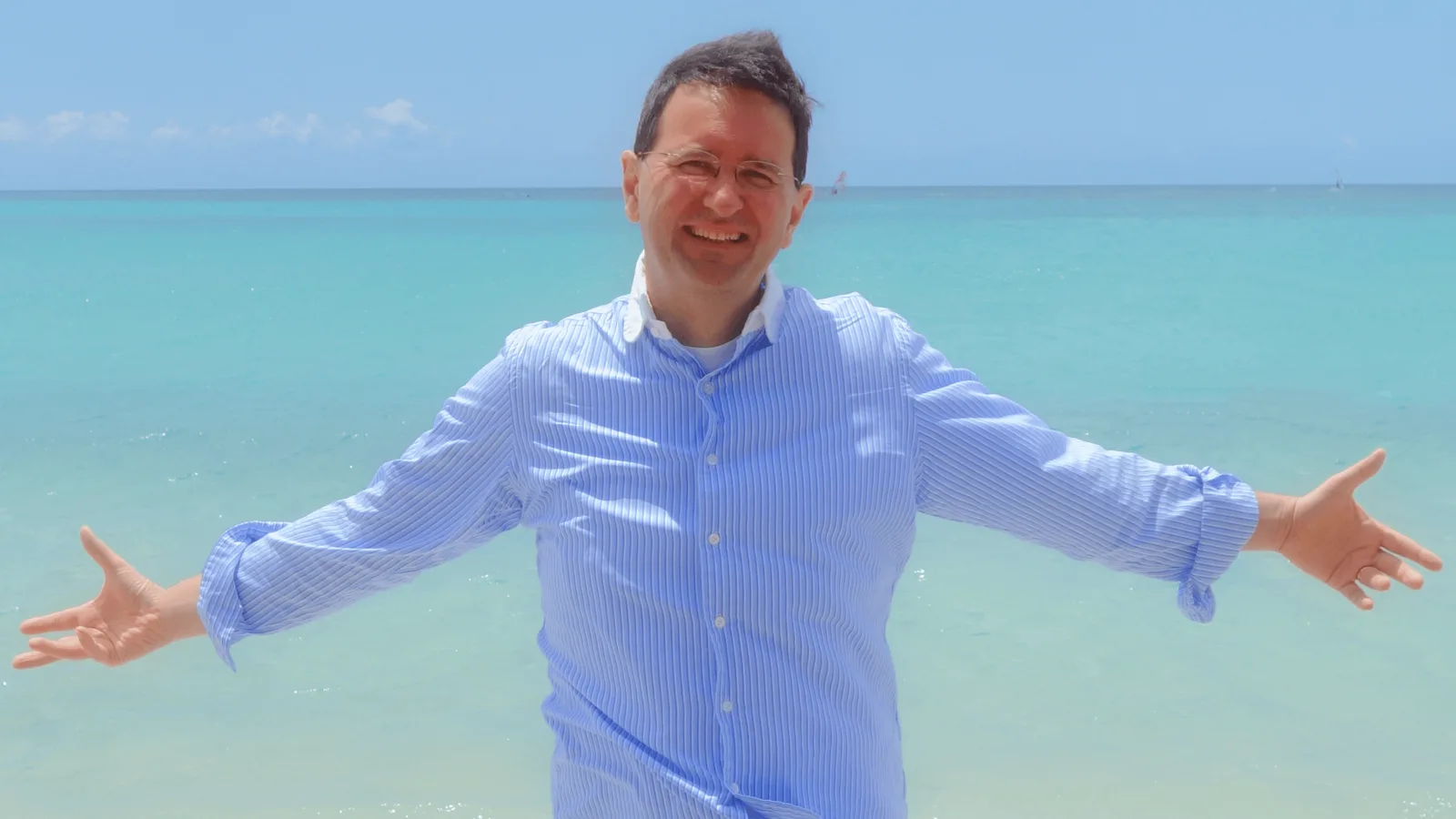 Mann in hellblauem Hemd mit ausgebreiteten Armen am Strand vor türkisblauem Meer und Himmel