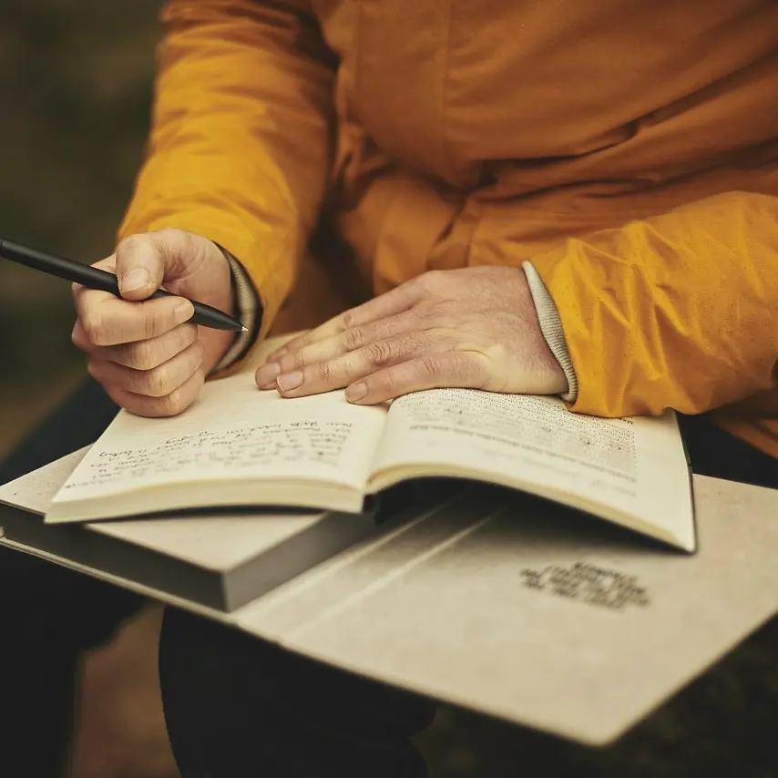 Person in gelber Jacke schreibt mit Stift in aufgeschlagenem Buch
