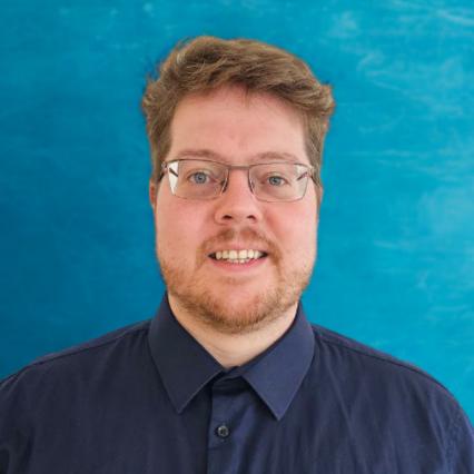Porträt von David Kölbach, Mann mit Brille vor blauem Hintergrund
