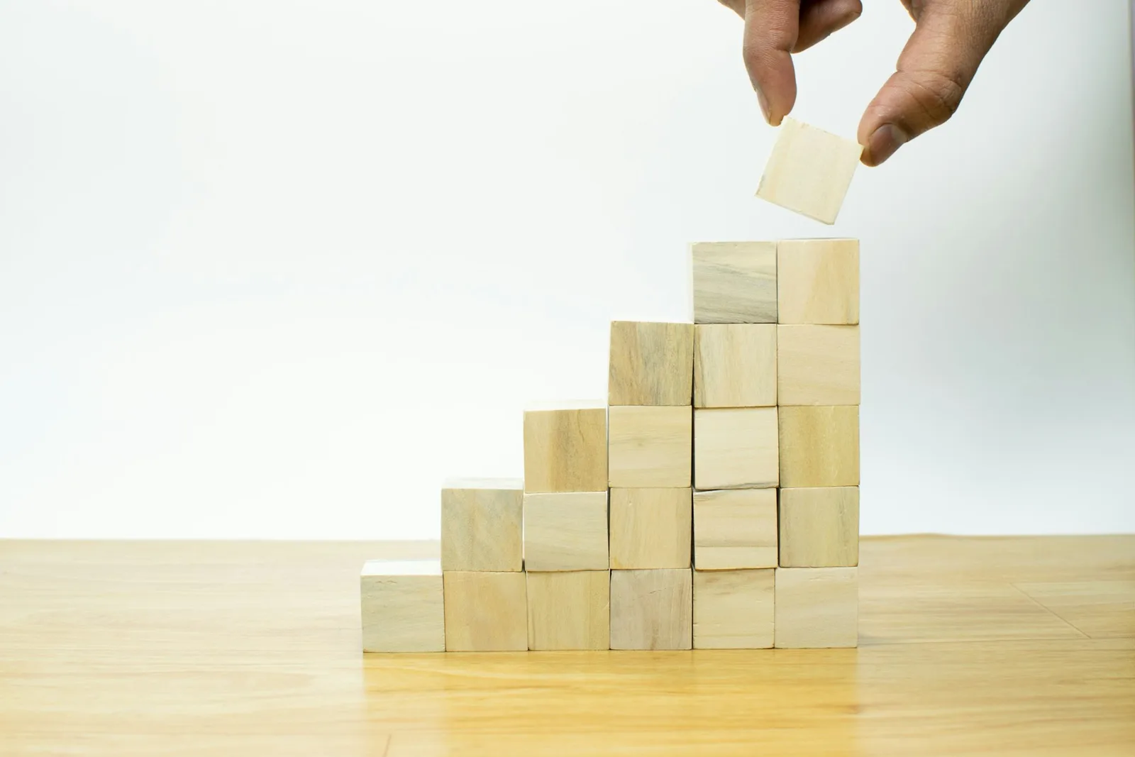 Hand setzt Holzwürfel auf Turm in Stufenform als Metapher für Aufbau und Progression