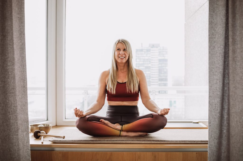 Nicole Freudenberg sitzt im Lotussitz in Yoga-Outfit vor Fenster mit Stadtblick