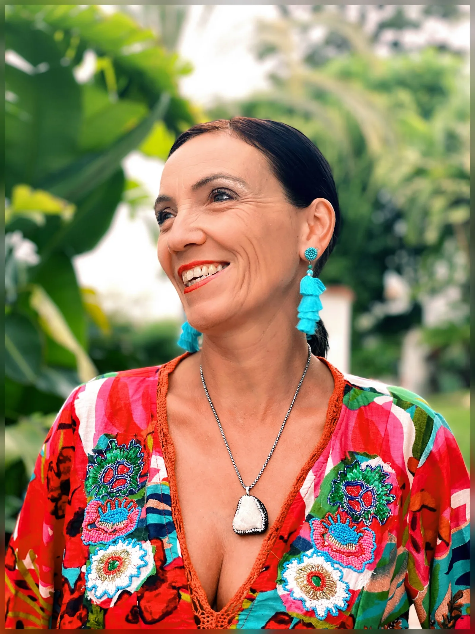 Carmen Duffner im bunten Patchwork-Kleid mit türkisen Ohrhängern, lachend vor Garten