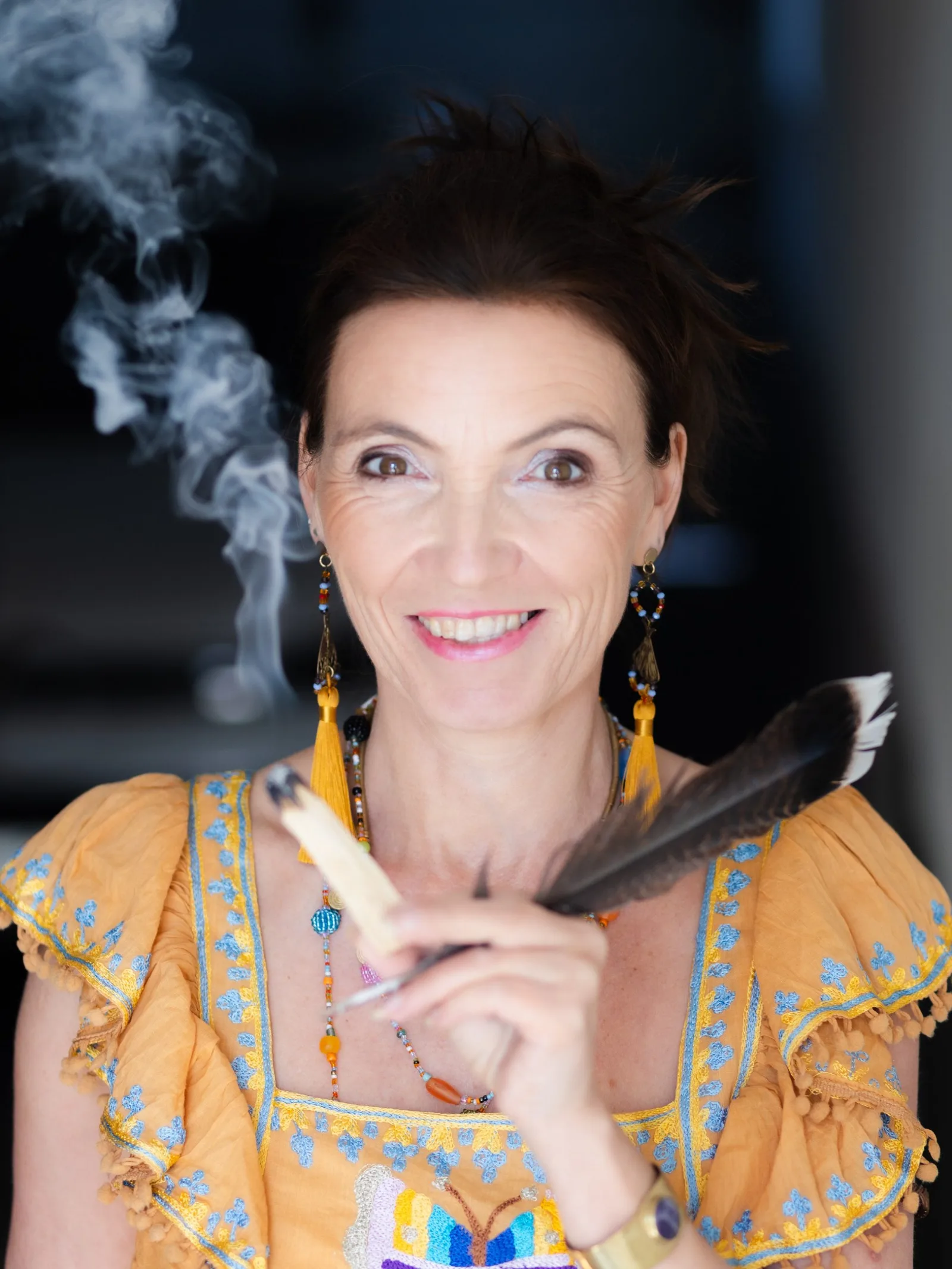 Carmen Duffner in gelbem Kleid hält Räucherstab vor dunklem Hintergrund, lächelnd