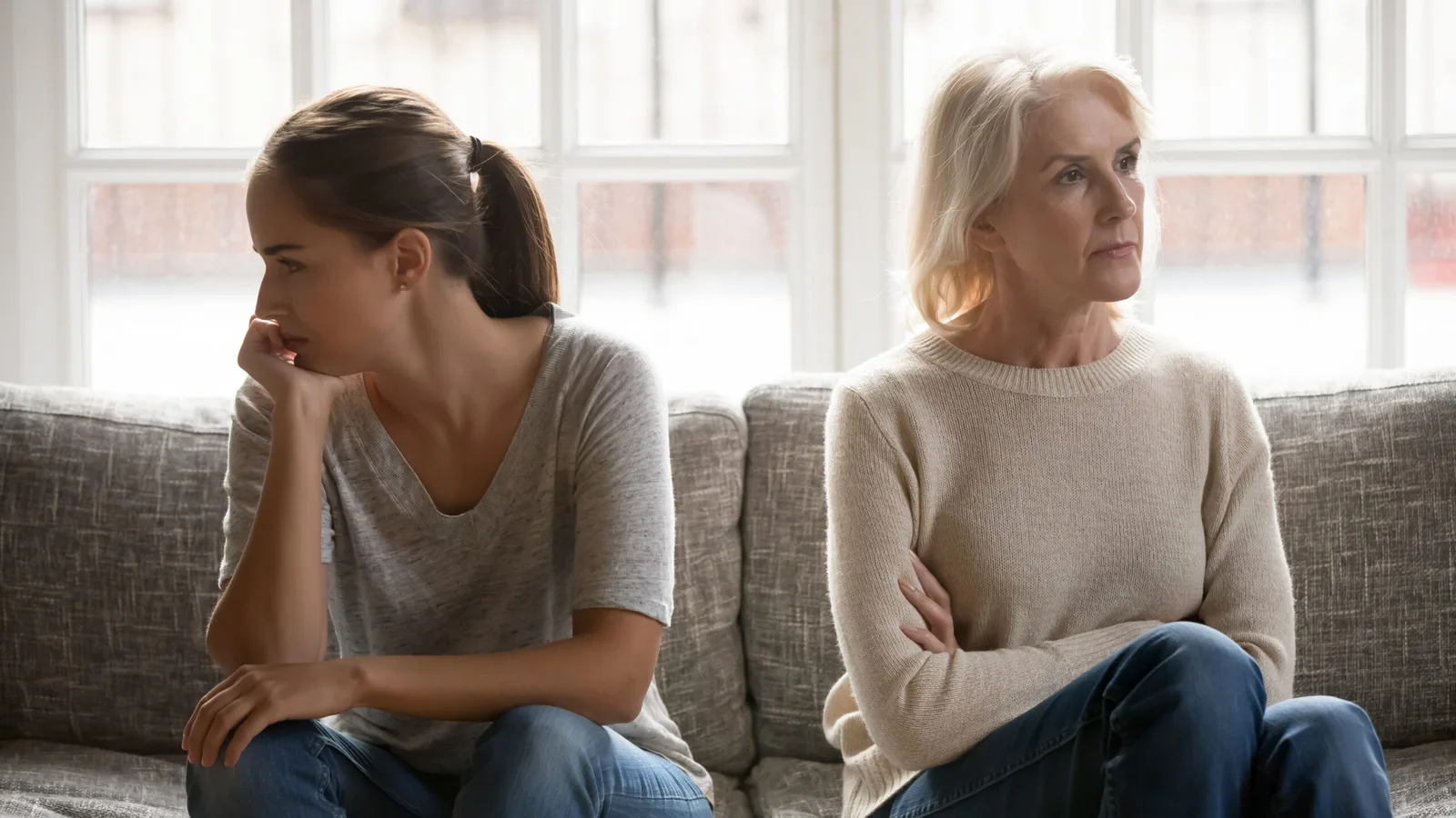 Zwei Frauen verschiedener Generationen auf Sofa, blicken besorgt und distanziert nebeneinander