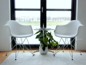 Modernes Therapie-Ambiente mit zwei weißen Designerstühlen, Glastisch und grüner Pflanze vor großem Fenster mit Stadtblick