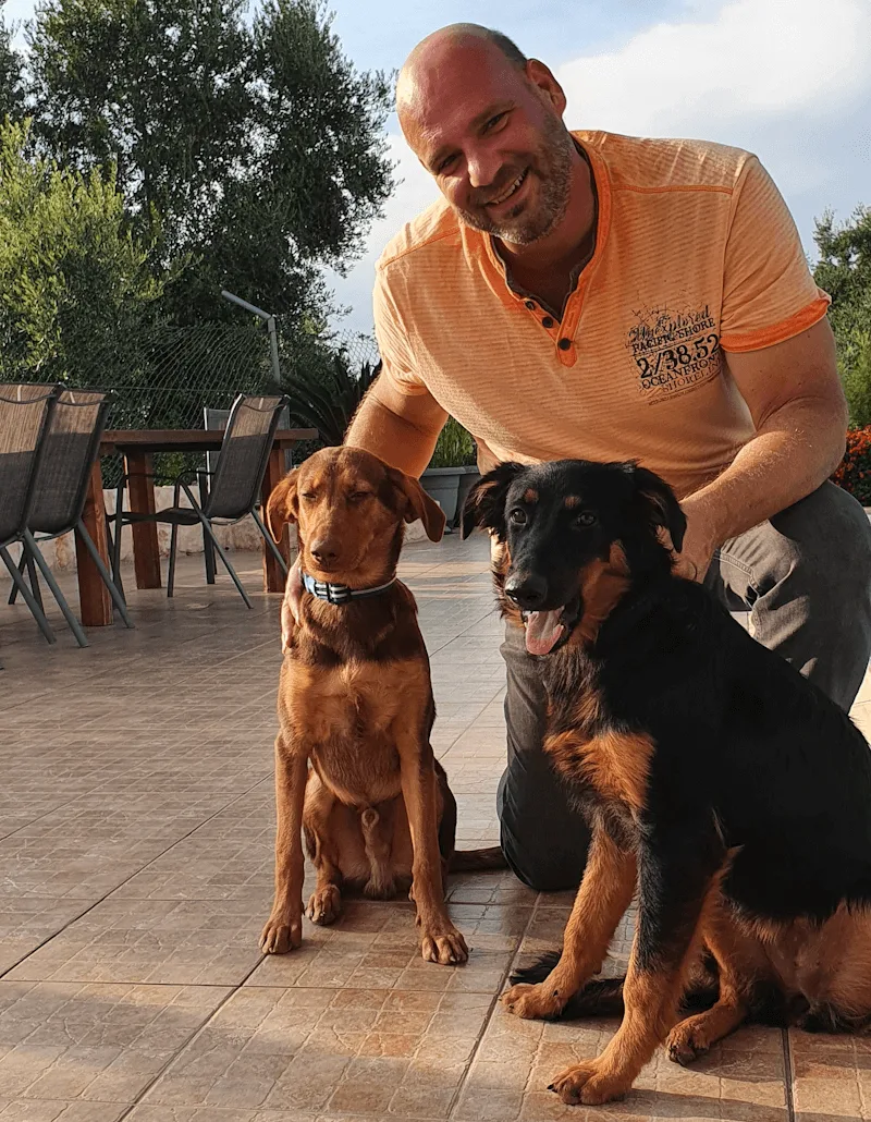 Jens Tell in orangem Polo-Shirt auf Terrasse mit zwei Hunden, braunem und schwarzbraunem Labrador