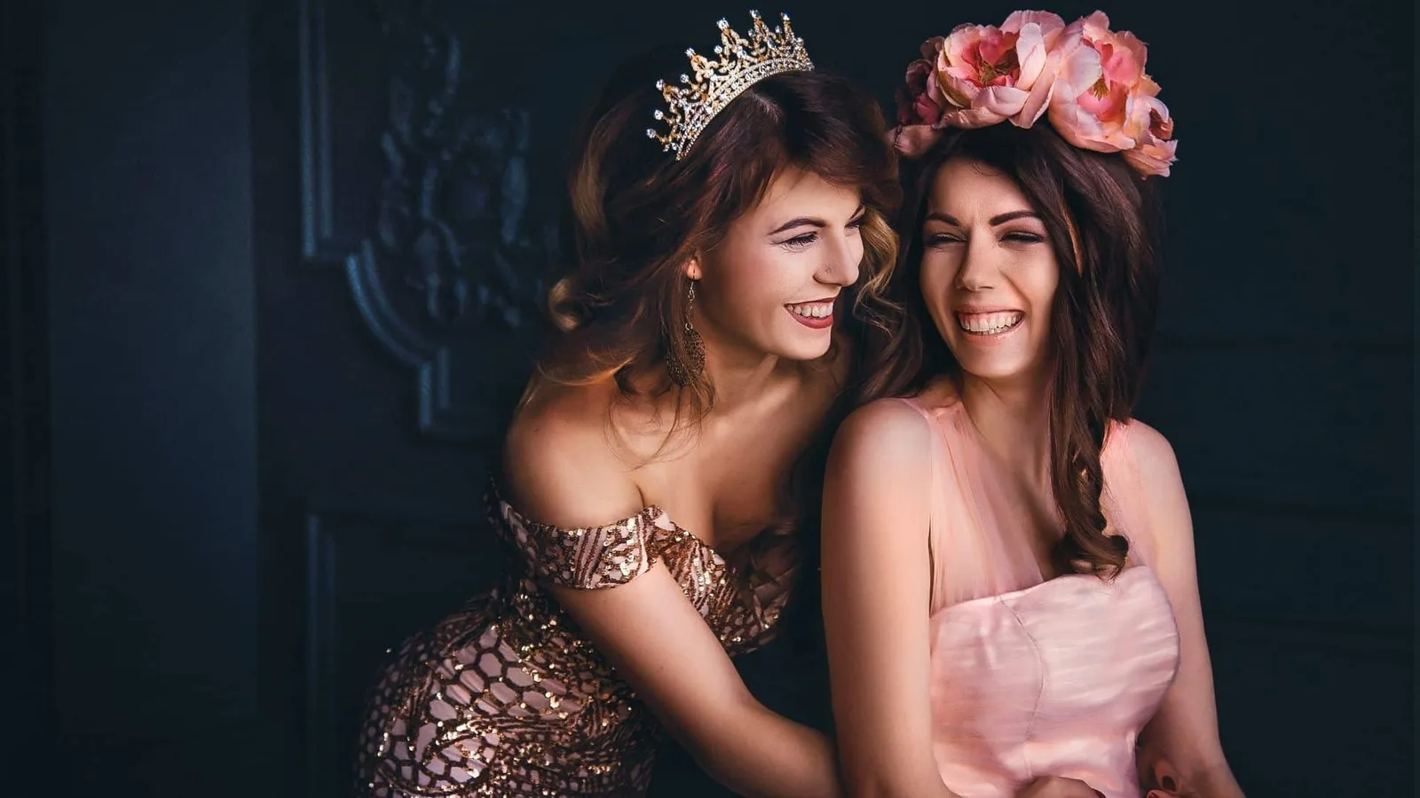 Zwei Frauen in eleganten Kleidern mit Krone und Blumenkranz, lachend vor dunklem Hintergrund