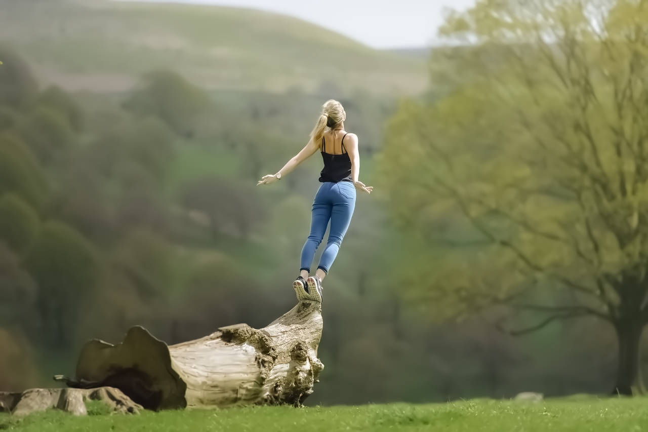 Frau springt über Baumstamm in grüner Naturlandschaft mit Bergen
