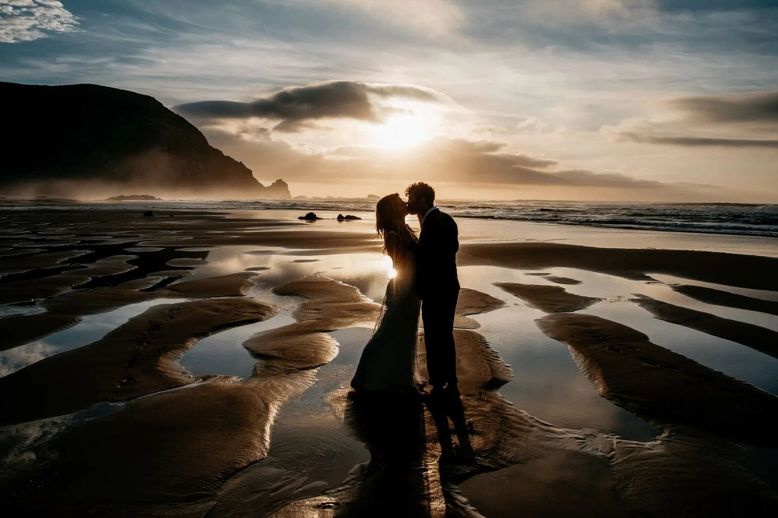 Couple kisses on beach with mountains at sunset, silhouetted against golden light
