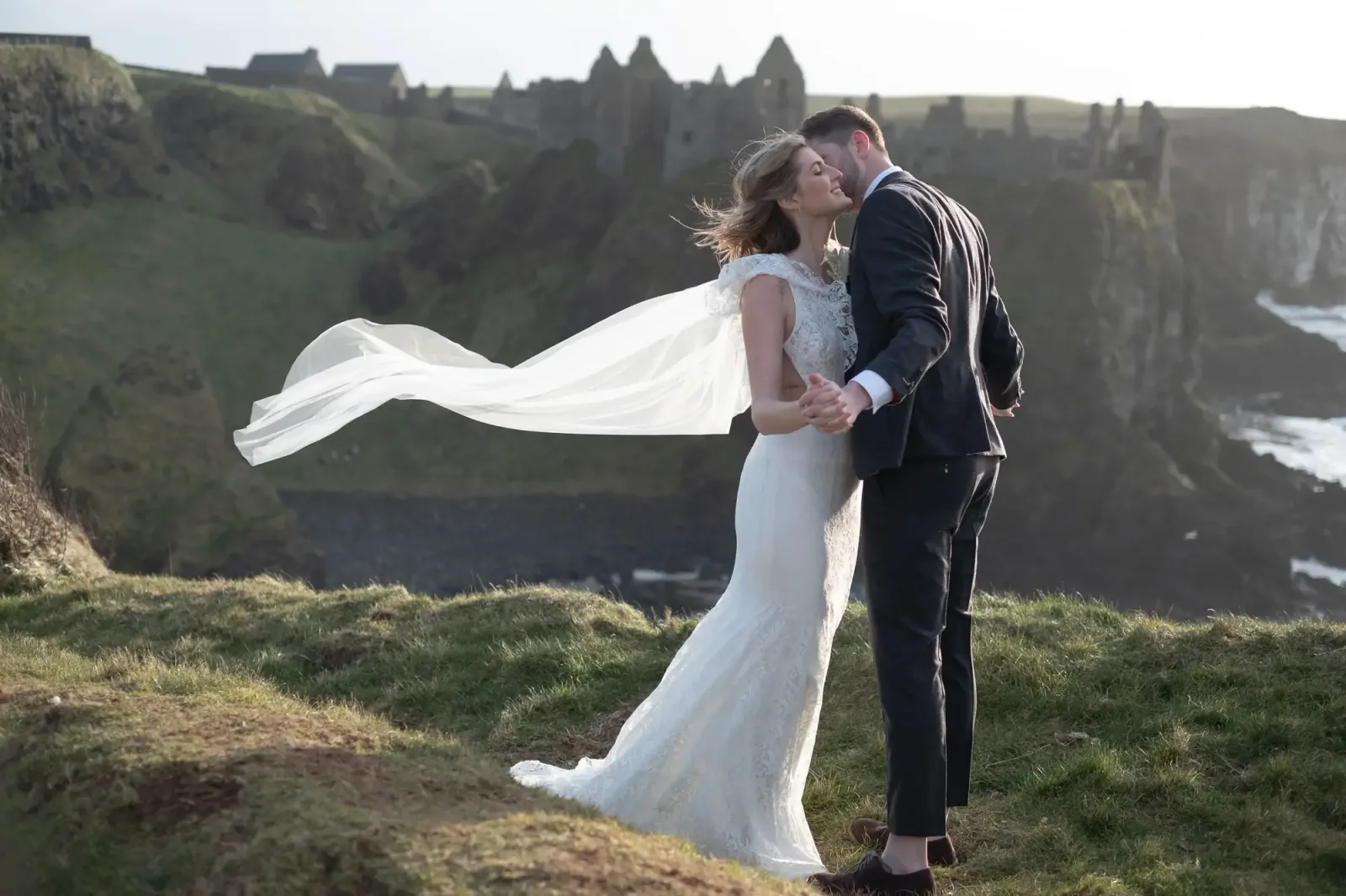 Brautpaar auf Klippenkante mit Burg im Hintergrund, fließendes Brautkleid, Hochzeitsfotografie