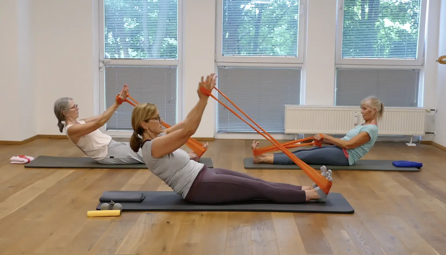 Drei Frauen auf Trainingsmatten, Pilates-Übung mit orangefarbenem Theraband für die Schultern