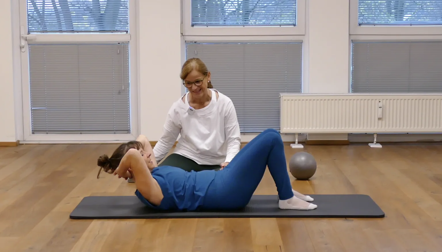 Trainerin führt Bauchmuskelübung durch, liegt auf schwarzer Matte mit Gymnastikball