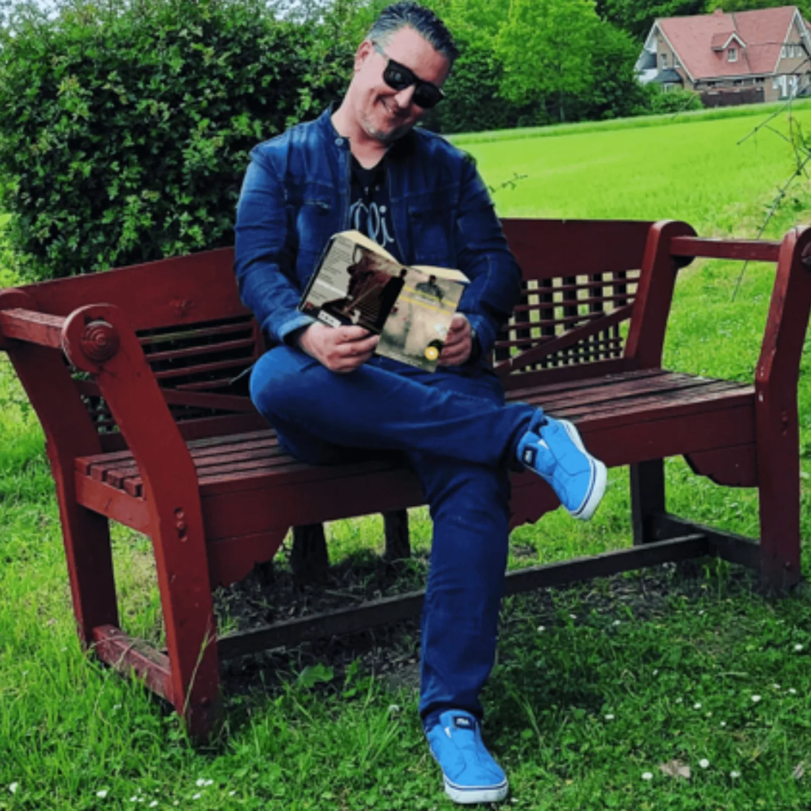 Hörbuchsprecher Ausbildung – Videokurs zum Stimmtraining 4 Christian Gera in Jeansjacke und Jeans sitzt auf einer roten Holzbank und liest ein Buch in einer grünen Landschaft.