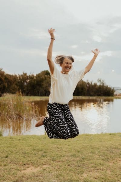 Frau beim Luftsprung in weißem Shirt und gemustertem Rock, glücklich lachend vor Wasser und Wiesen