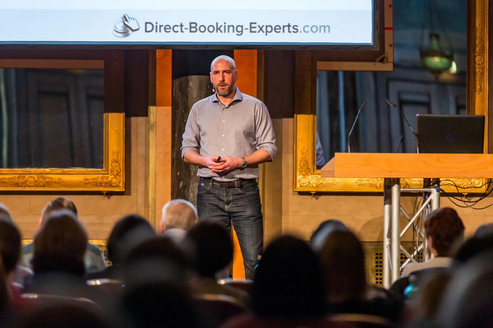 Redner auf Bühne vor Publikum mit Direct-Booking-Experts Logo
