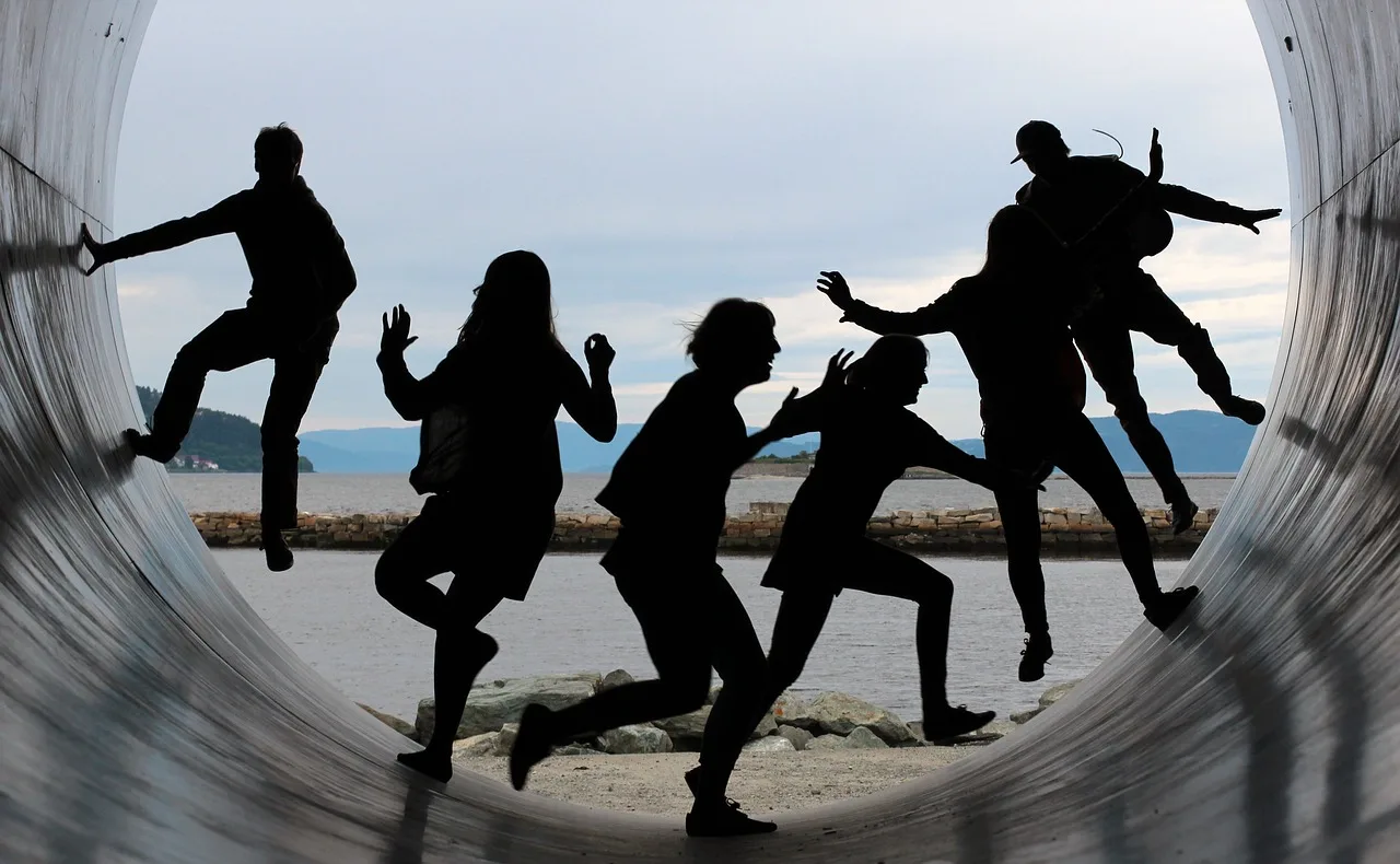 Silhouetten von fünf Menschen in dynamischen Posen, die durch ein großes Rohr springen, mit Meer und Bergen im Hintergrund