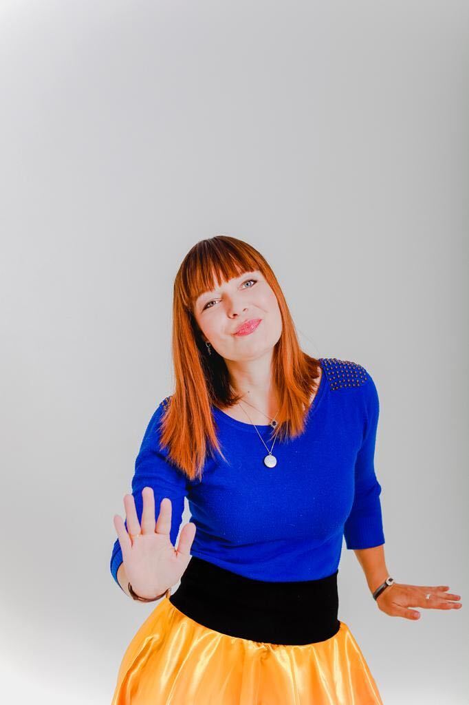 Portrait-Fotografie einer Frau mit roten Haaren und Pony in blauem Pullover und gelbem Rock vor grauem Hintergrund