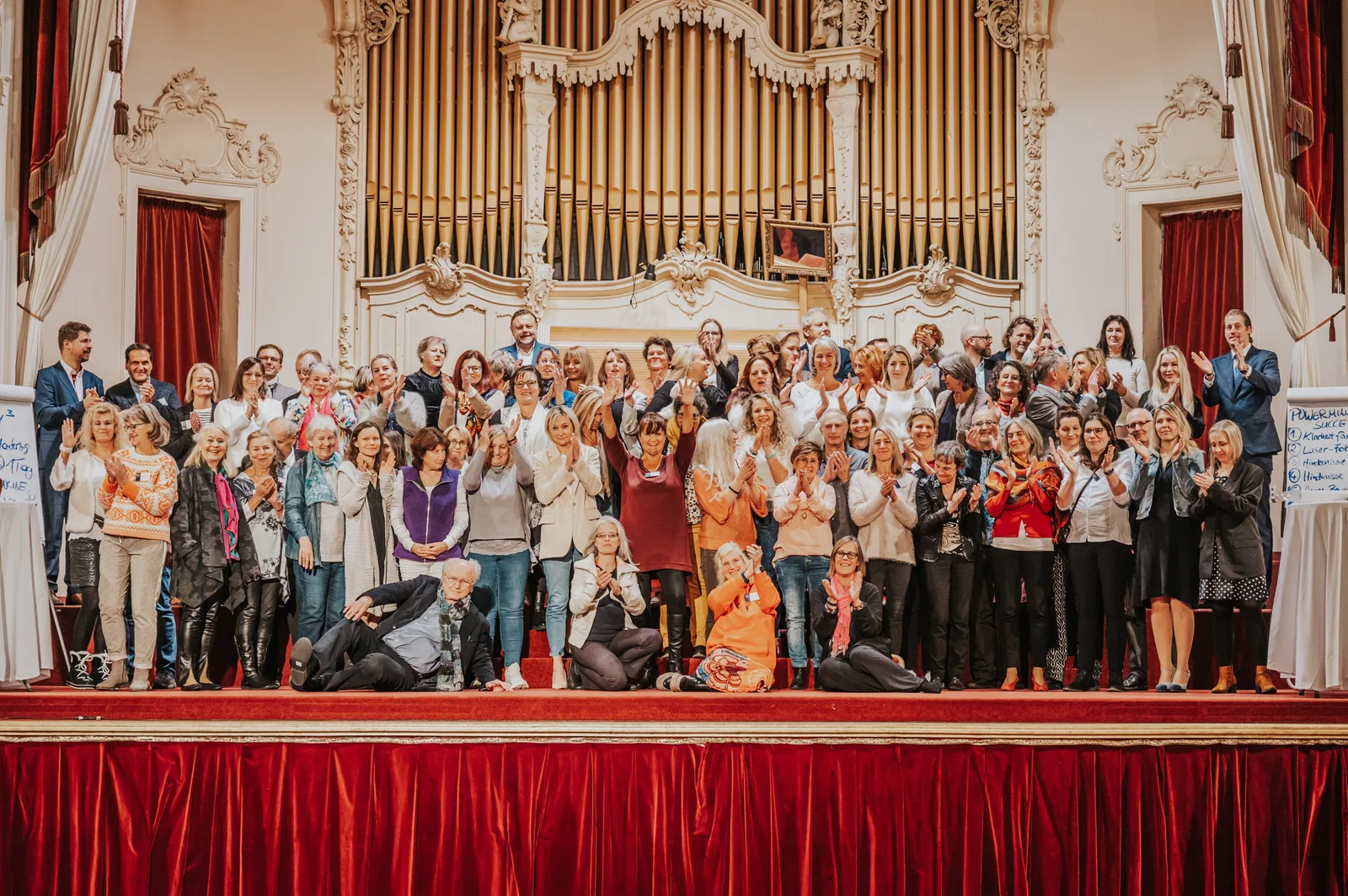 Große Gruppe Teilnehmer applaudiert auf Veranstaltungsbühne vor Orgel