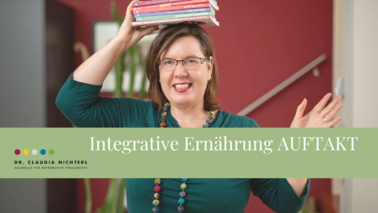 Dr. Claudia Nichterl hält bunte Bücher in die Luft, trägt grüne Bluse und Brille vor rotem Hintergrund