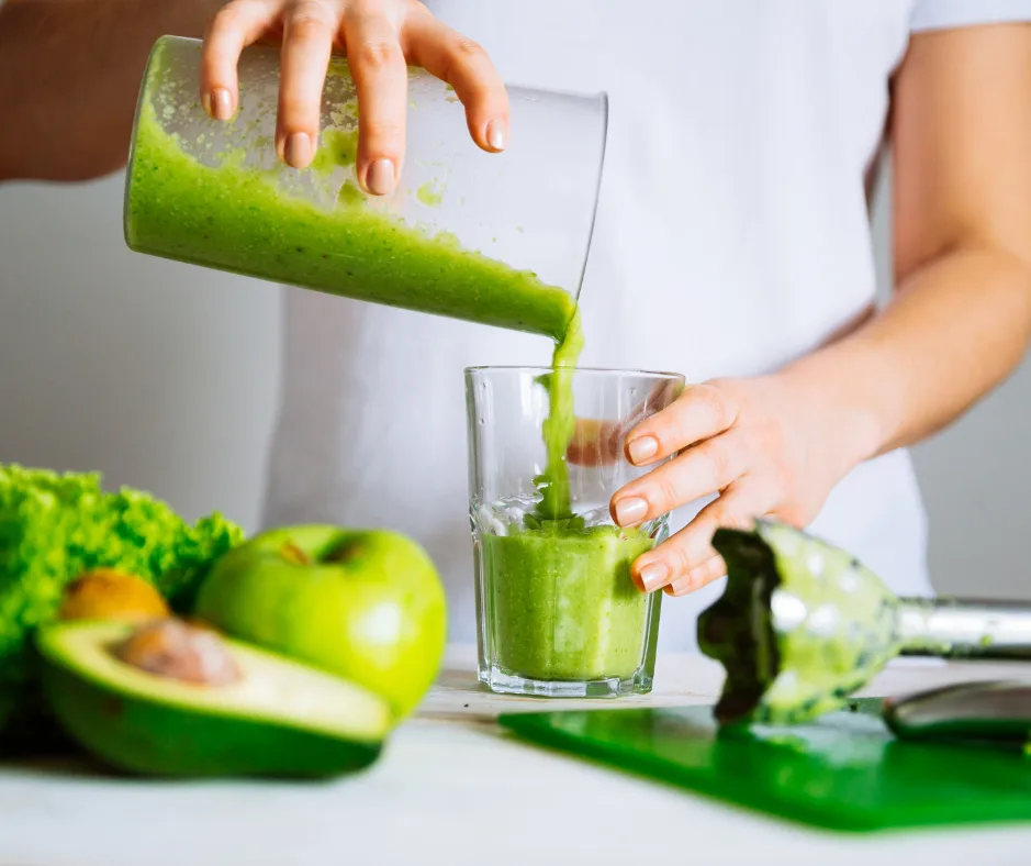 Hand gießt grünen Smoothie aus Mixer in Glas mit Avocado und Apfel im Hintergrund