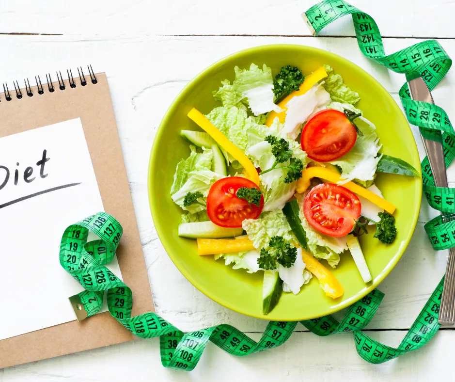 Grüner Teller mit frischem Salat, Tomaten, Paprika, Gurke neben Notizbuch mit "Diet" beschriftet und grünem Maßstab
