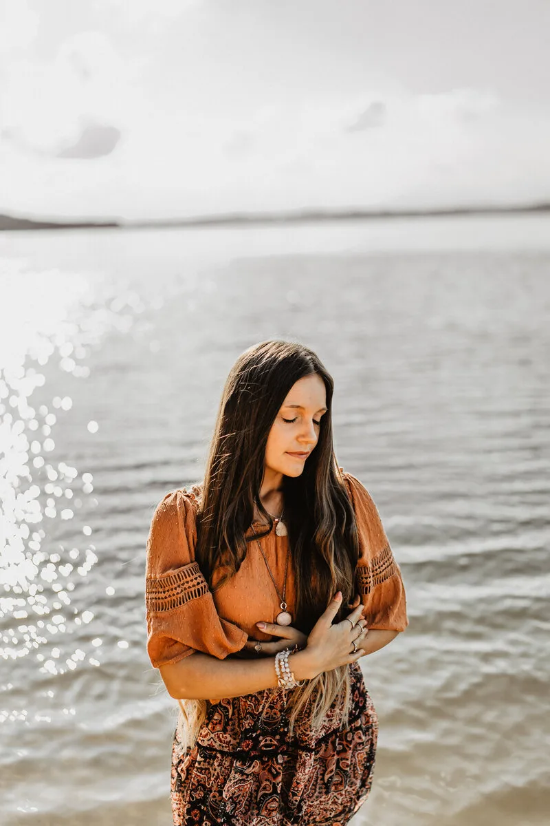 Intuitionsguide – Videokurs zur Selbstbefreiung 3 Sarah Jane Rogalski in orangebrauner Bluse am Wasser stehend, nach unten blickend, Sonnenlicht auf dem See