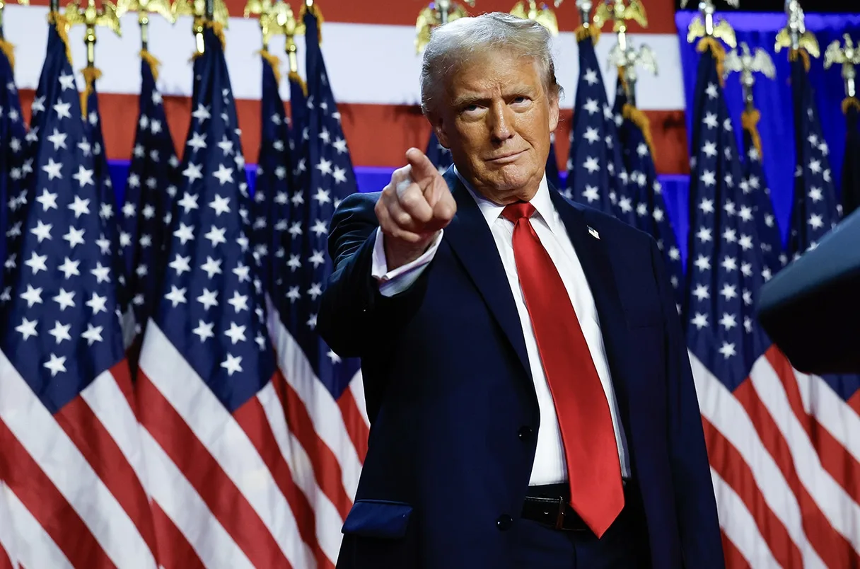 Ein Mann im Anzug mit roter Krawatte zeigt nach vorne und verkörpert den politischen Hype. Er steht vor mehreren amerikanischen Flaggen und einem rot-weiß-blauen Hintergrund, was an den Rummel rund um das Phänomen der Trump Meme Coin erinnert.
