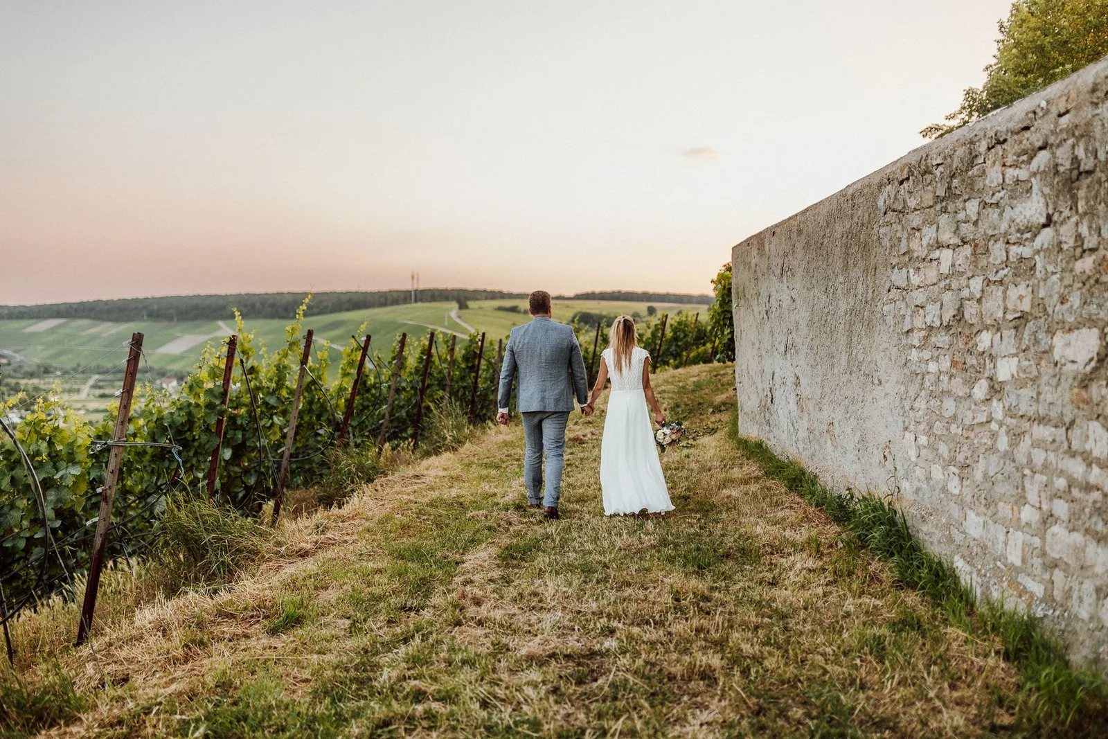 Brautpaar spaziert Hand in Hand durch Weinberge bei Sonnenuntergang mit Steinmauer
