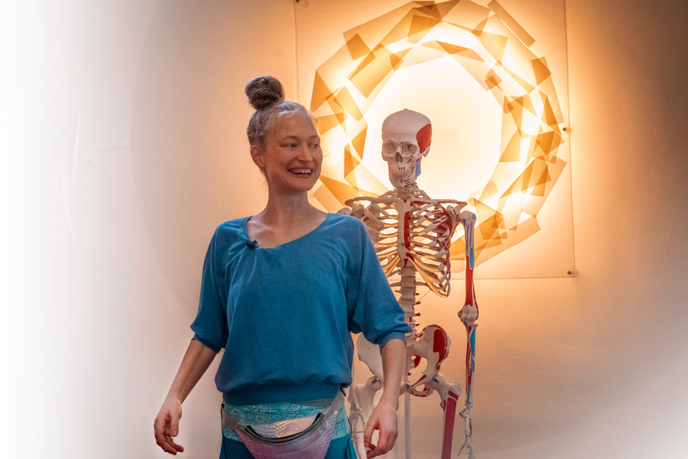 Frau lächelt neben anatomischem Skelettmodell vor goldenem Licht-Mandala im Hintergrund