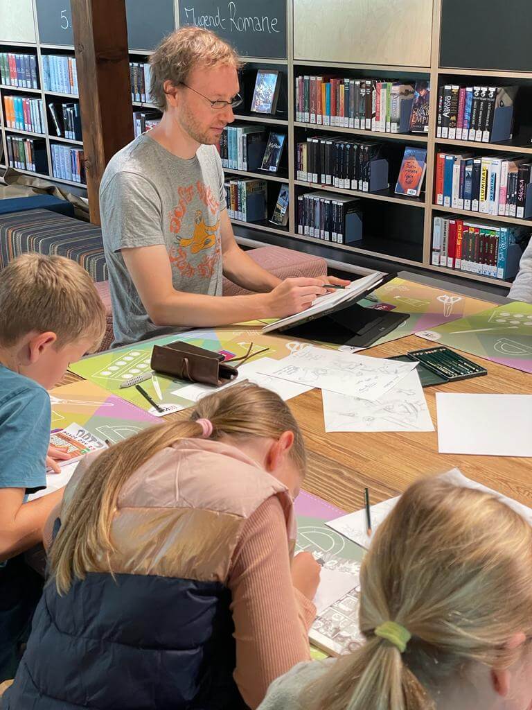 Maxim unterrichtet Kinder beim Manga-Zeichnen an Tisch mit Materialien