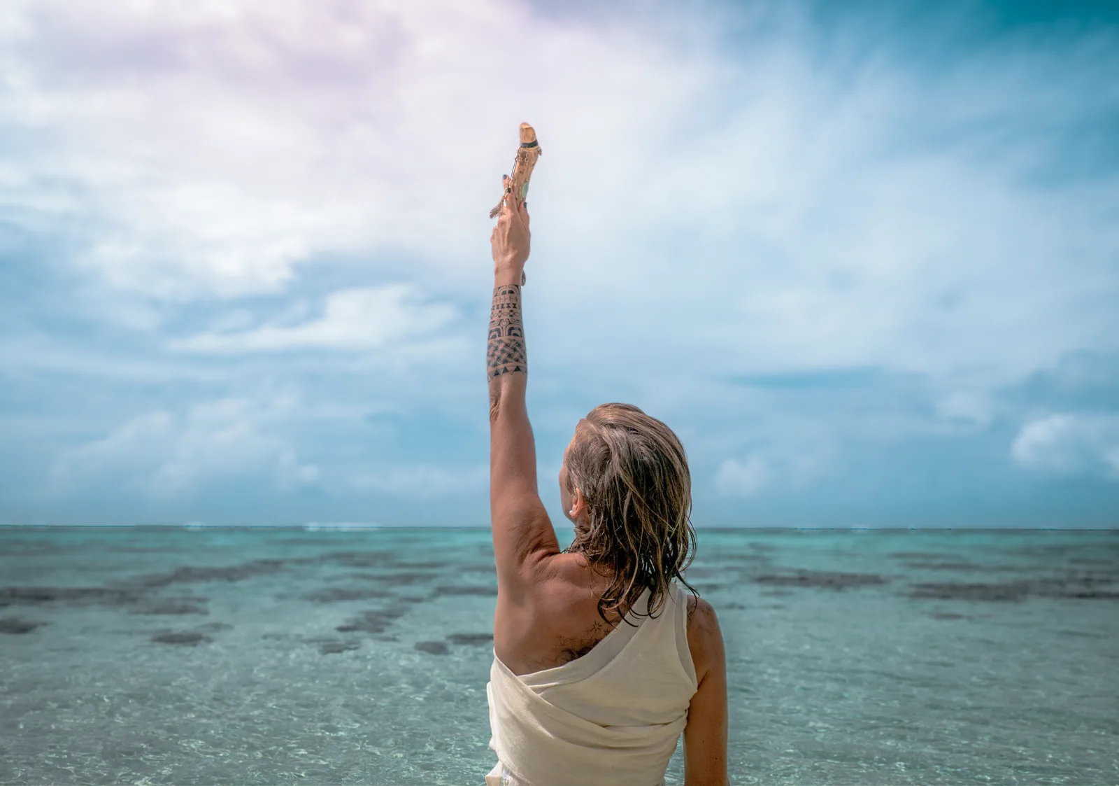 Kaimana und die 12 Schlüssel – Coaching zur Selbsterkenntnis 2 Frau mit erhobenem Arm am Meer, Blick zur Weite, weißes Oberteil, türkises Wasser und Himmel