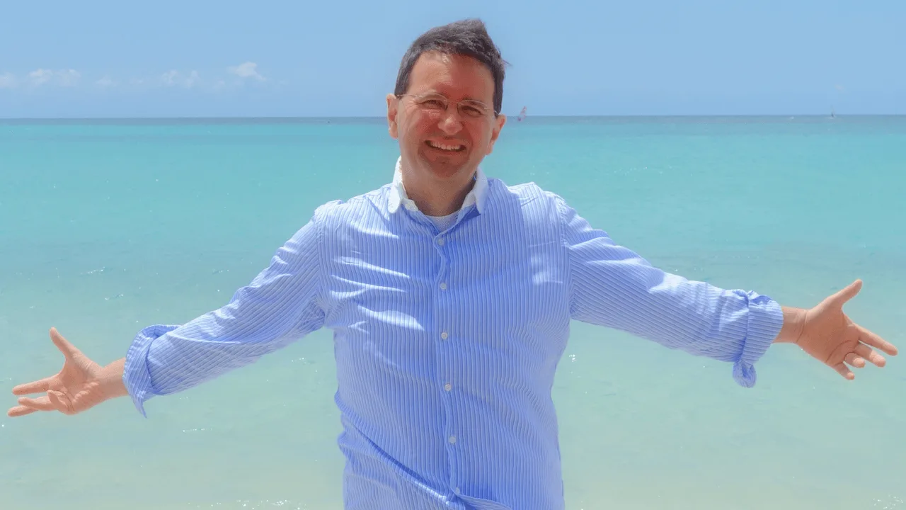 André Loibl am Strand mit blauem Hemd und ausgebreiteten Armen vor türkisem Meer