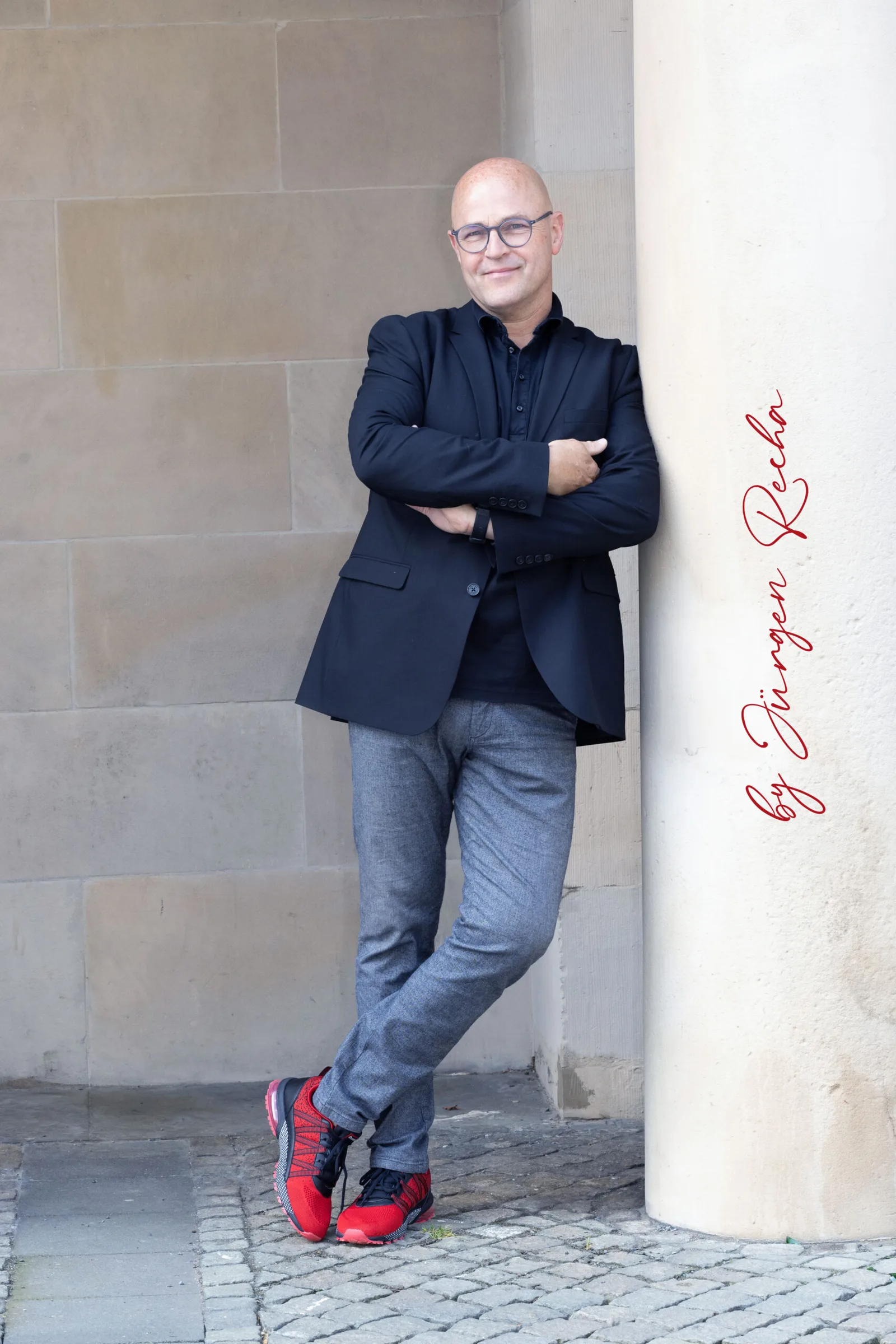 Porträt von Jürgen Recha, Mann mit Brille in dunklem Blazer und Jeans, lehnt an Säule mit roten Sneakern