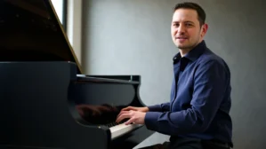 Roman Sillipp sitzt am Flügel im Studio, trägt blaues Hemd, lächelt zur Seite