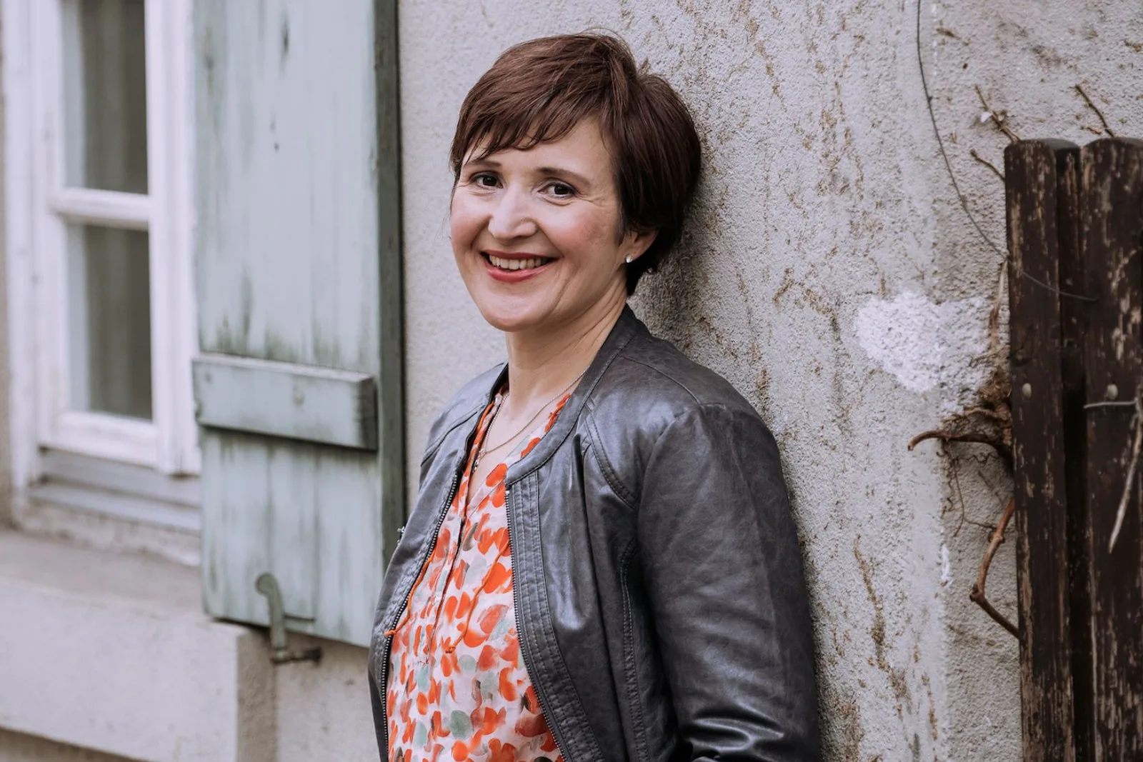 Porträt Gabriele Feile, braune kurze Haare, rotes Muster, schwarze Jacke, lächelnd, Hauswand, ganzer Körper