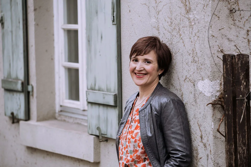 Porträt Gabriele Feile, braune kurze Haare, oranges Muster, schwarze Jacke, lächelnd vor grauer Hauswand