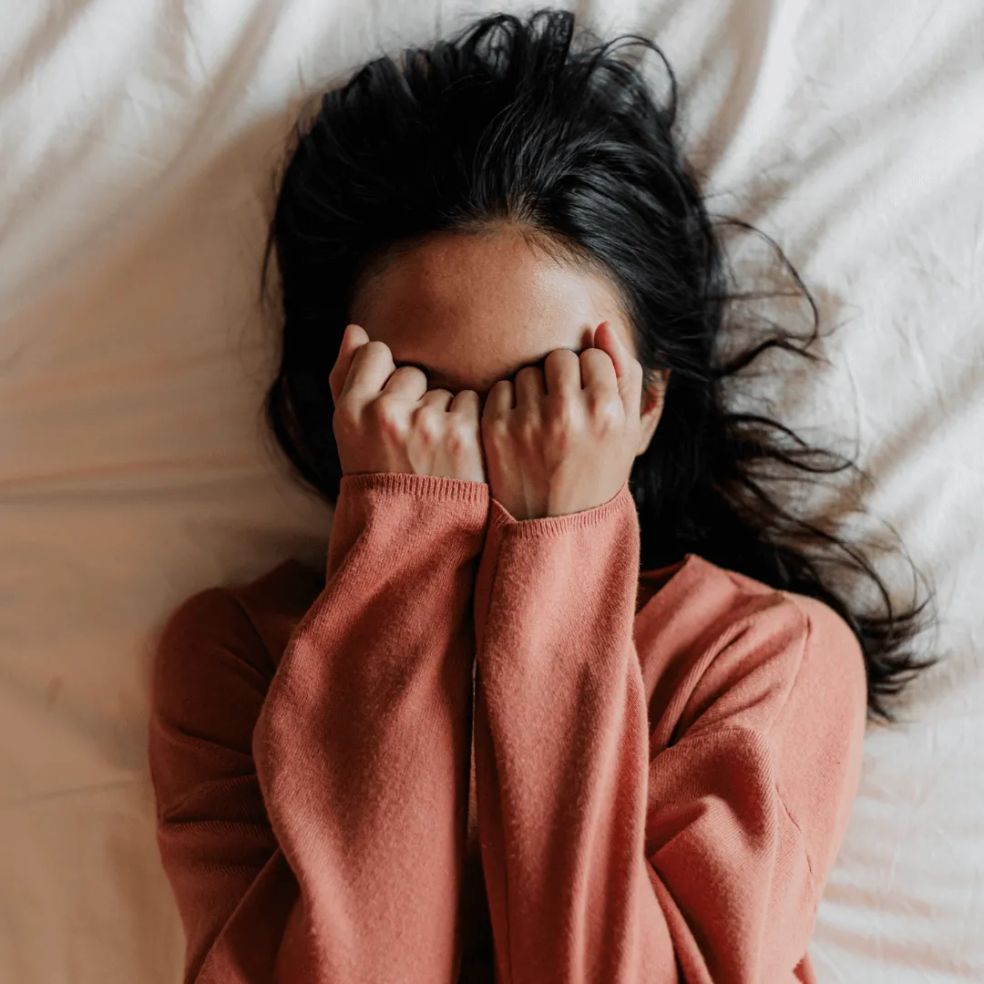 Klopf dich in deine Mitte: EFT – Videokurs zur Stressregulation 3 Frau mit dunklem Haar sitzt überfordert auf weißem Bett, verdeckt Gesicht mit Händen in rosafarbener Kleidung