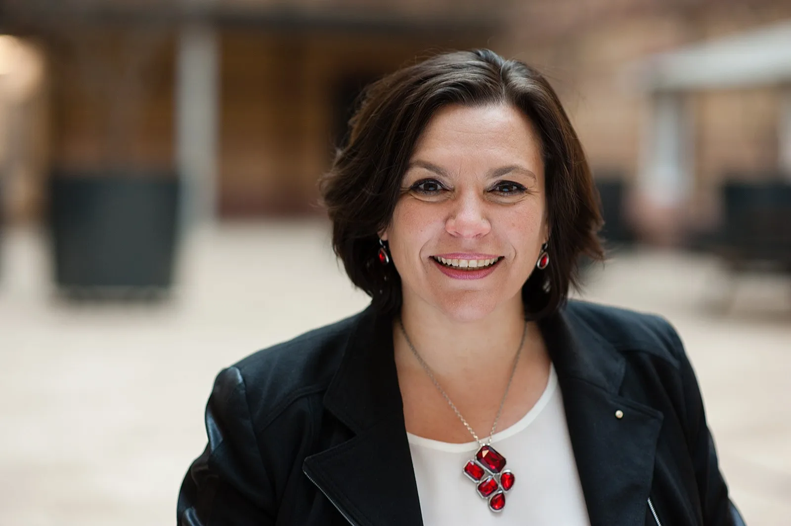 Christine Hofmann Portrait im schwarzen Blazer mit rotem Schmuck, lächelnd, Büro-Hintergrund, professionell