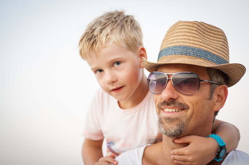 Kind auf Schultern von Mann mit Hut und Sonnenbrille vor weißem Hintergrund