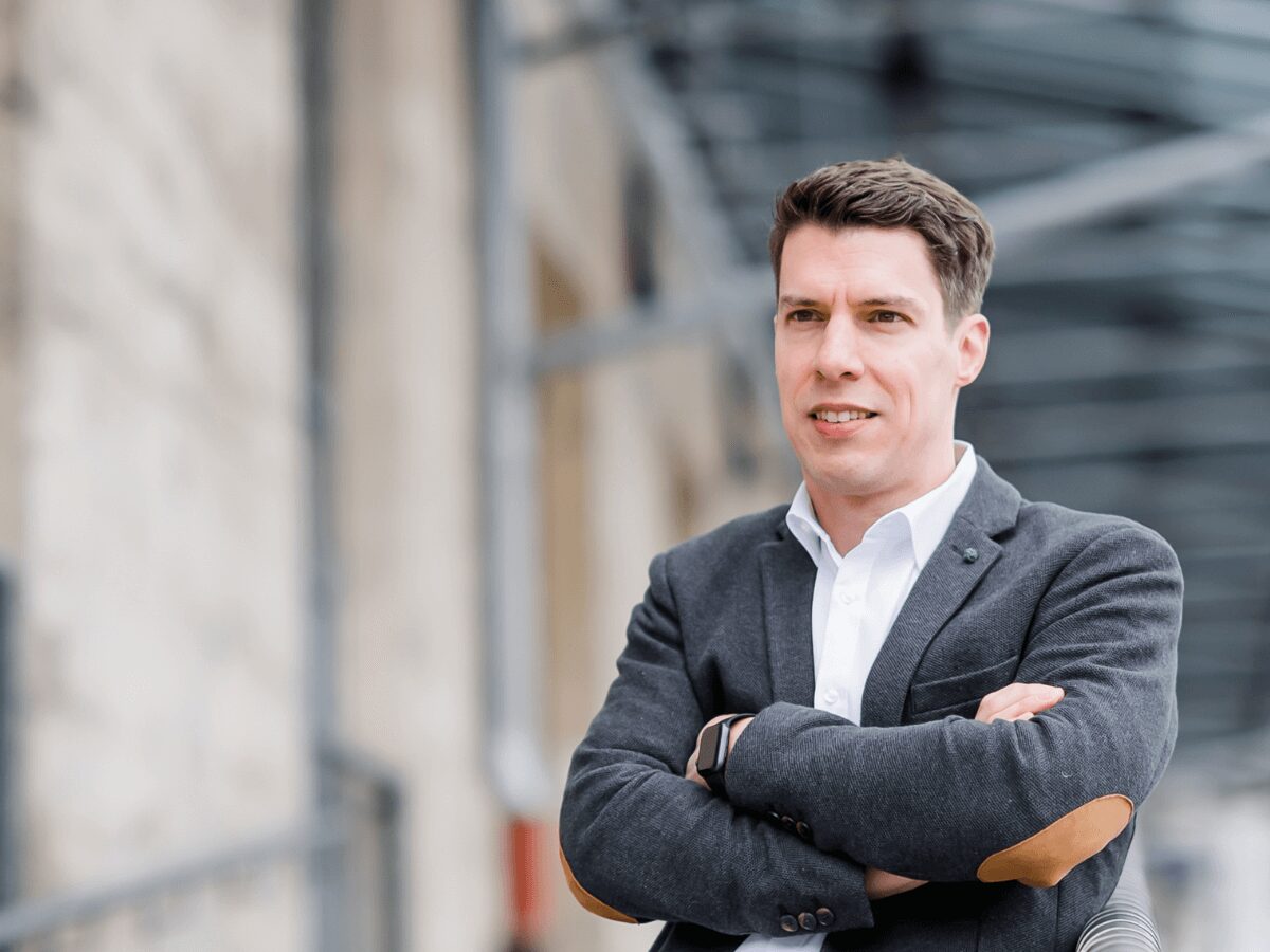Mann im grauen Blazer mit verschränkten Armen vor unscharfem Hintergrund, Einzeltraining Konflikte souverän meistern