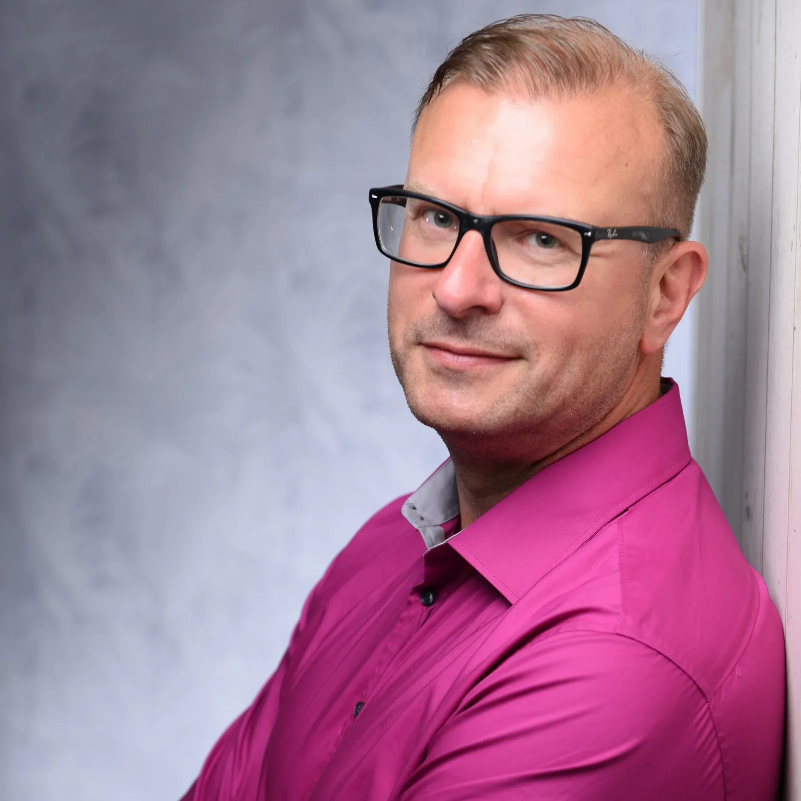 Porträt von Andreas Dannebauer mit Brille und pink gefärbtem Shirt vor grauem Hintergrund