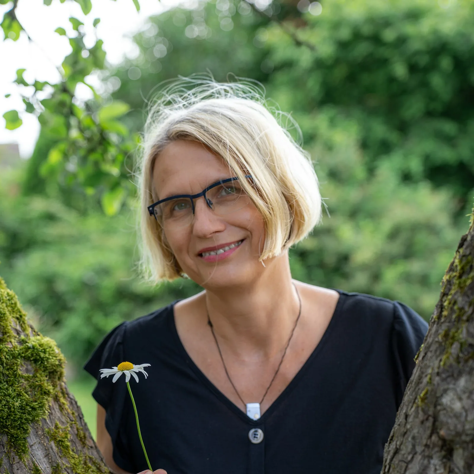 Porträt von Kathleen Kühn mit blondem Haar, Brille, schwarzem Shirt mit Gänseblümchen im grünen Hintergrund