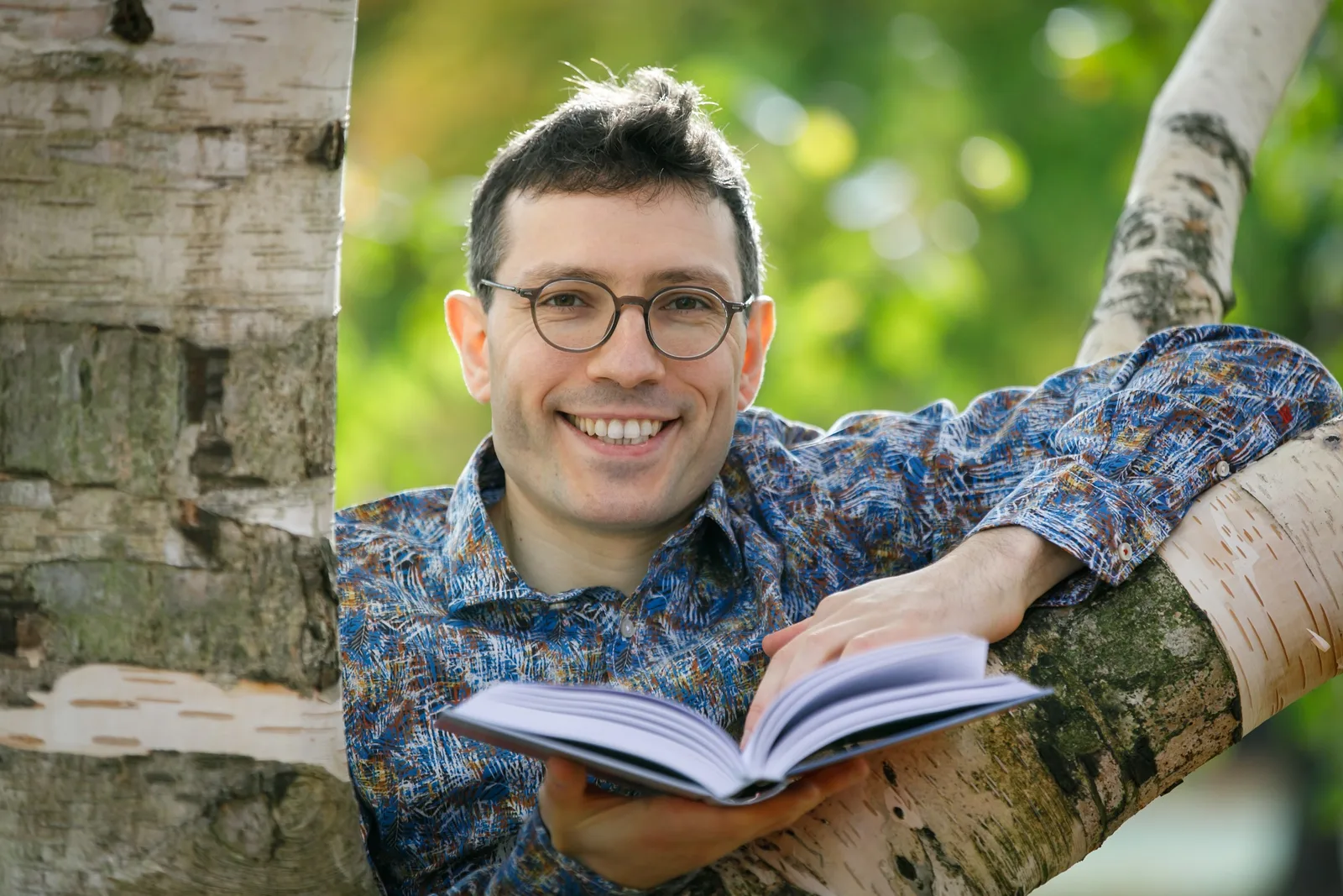 Evgenij Unker sitzt an Birkenbaum mit aufgeschlagenem Buch, Naturhintergrund