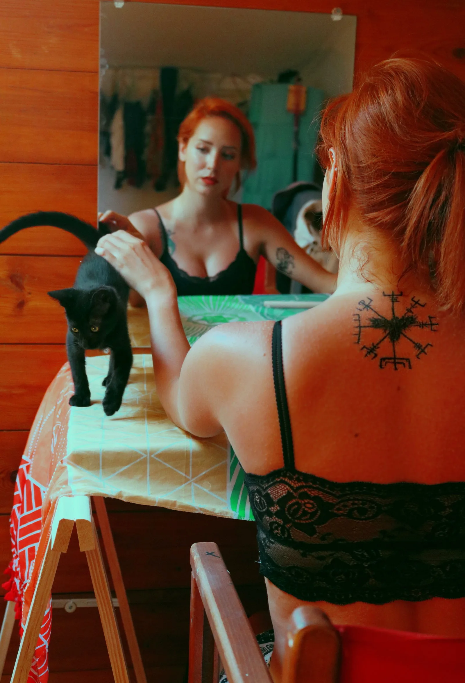 Rothaarige Frau vor Spiegel mit Runen-Tattoo auf Rücken und schwarzer Katze