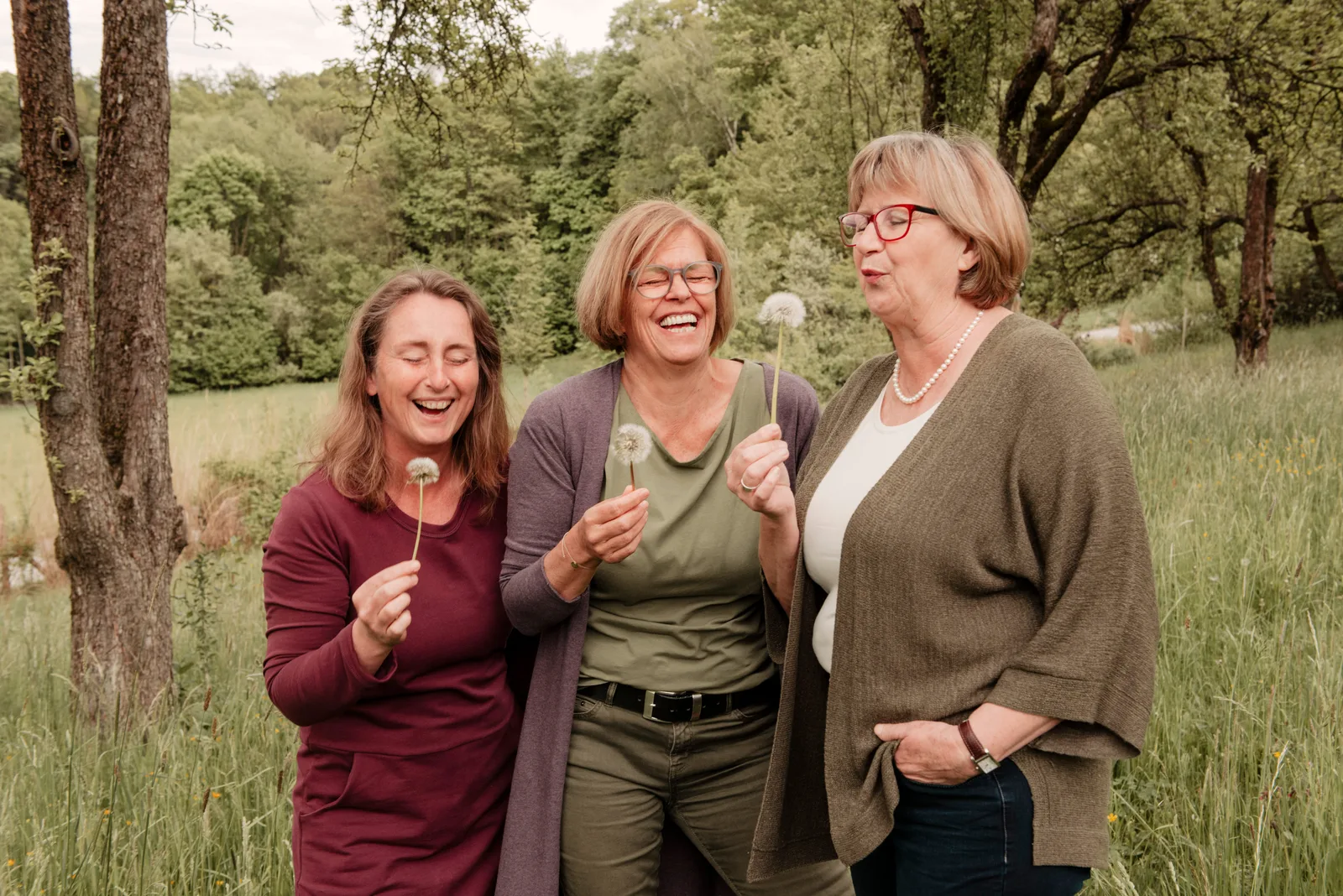 Drei Frauen in Freizeitkleidung lachend in einer Waldwiese, halten Pusteblumen, Team-Foto